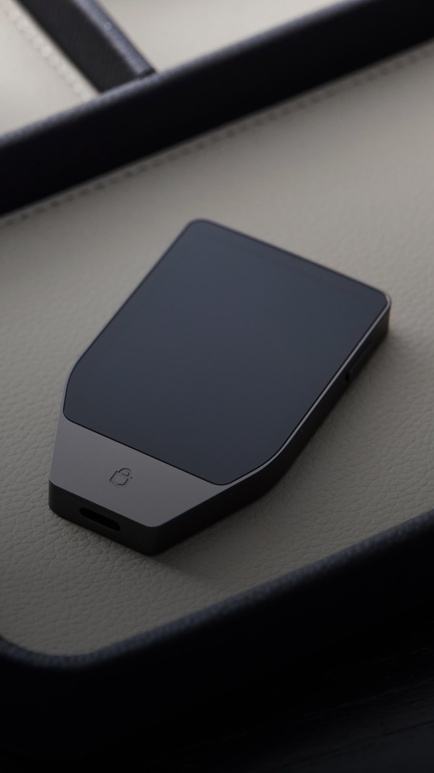 A man sits on a chair using a MacBook while holding a Trezor Safe 7 hardware wallet in one hand.