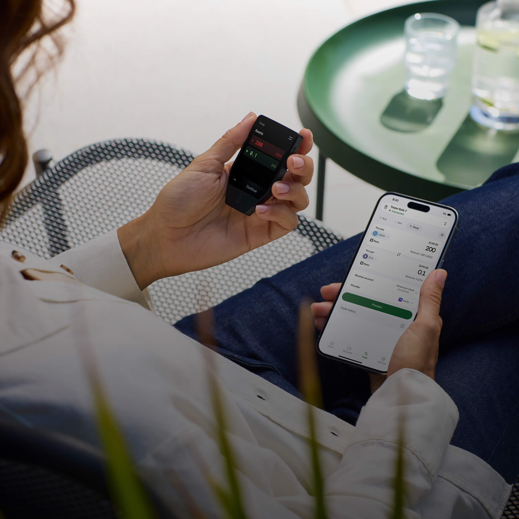 Blurry rear view of a man holding a smartphone in his left hand and a Trezor Safe 7 hardware wallet in his right hand.