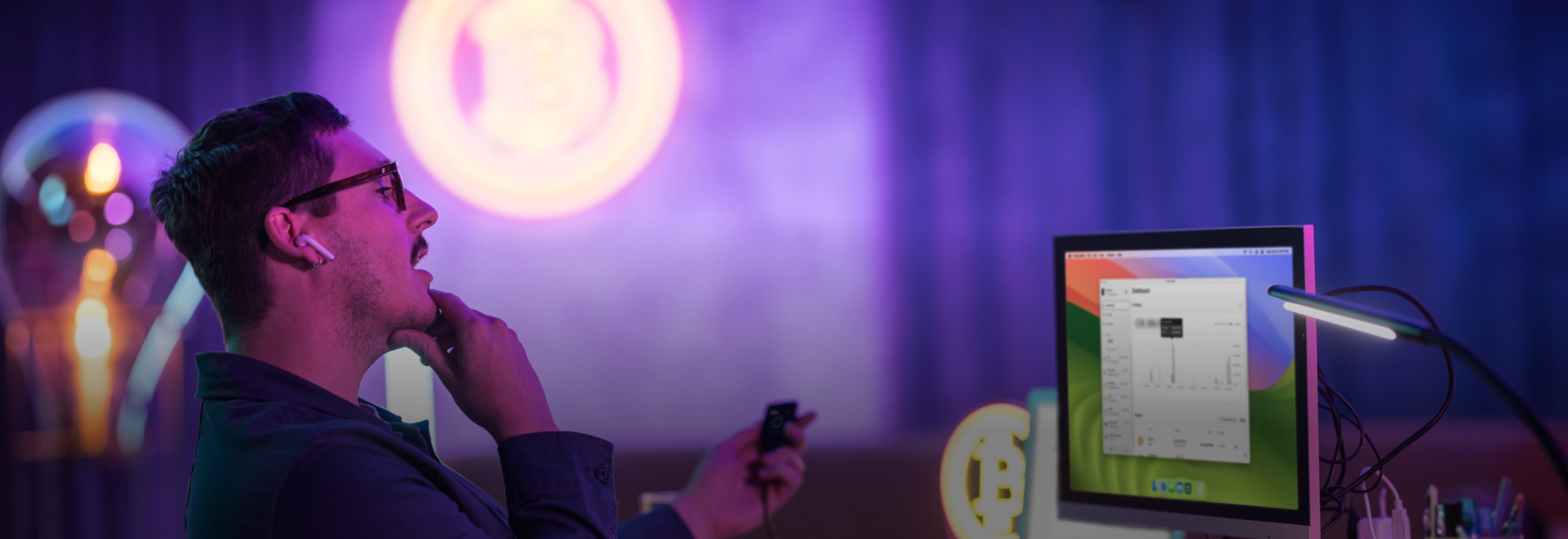 A man wearing glasses and wireless earbuds sits at a desk, looking thoughtfully at a computer monitor showing a Trezor Suite dashboard. He's holding a Trezor Safe 5 in one hand, resting his chin on the other. The background features neon Bitcoin symbols and a dimly lit room with purple and blue lighting for a modern, tech-focused feel.